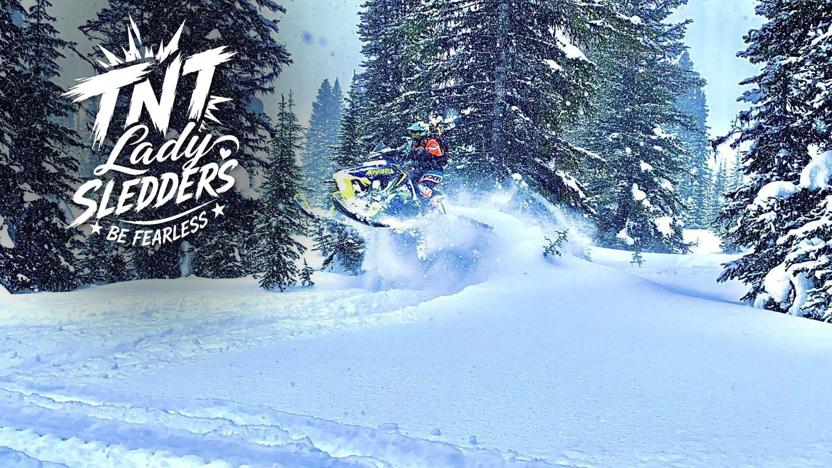 TNT Lady Sledders logo over Gisele riding through deep powder between snow-covered pines